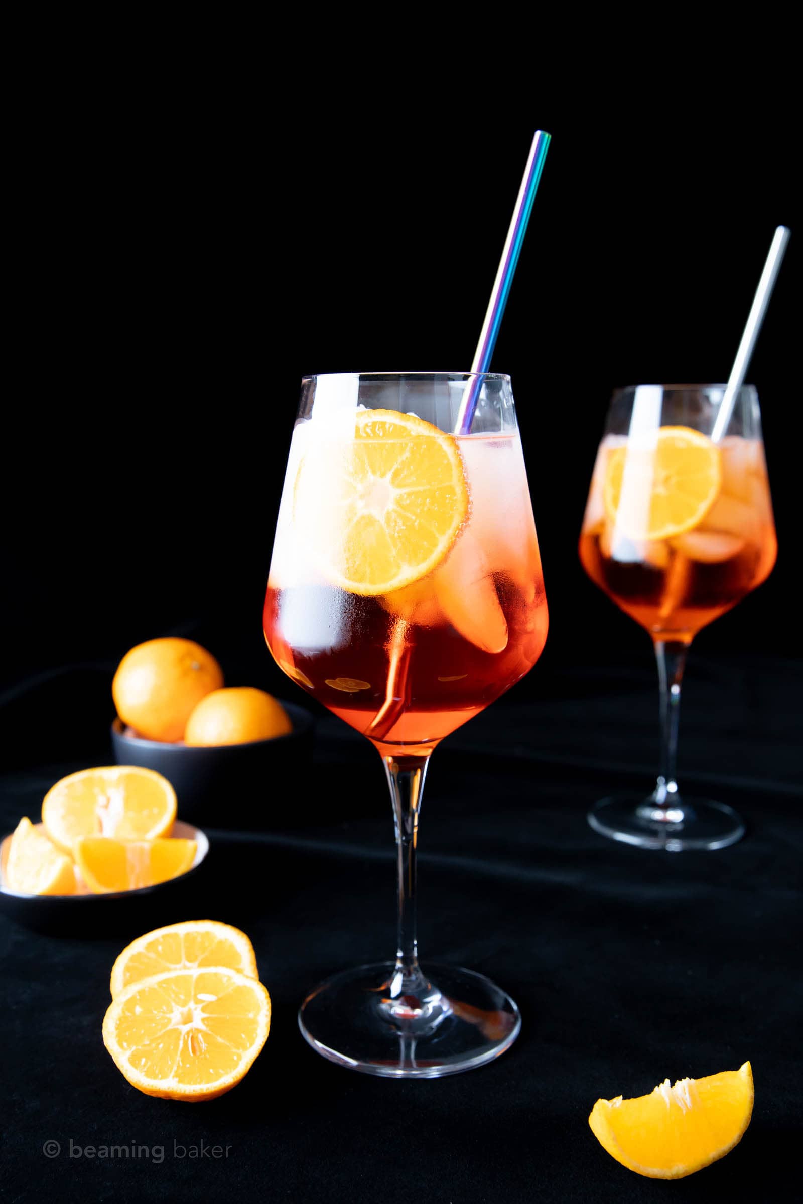 two wine glasses full of Aperol Spritz against a black backdrop with oranges