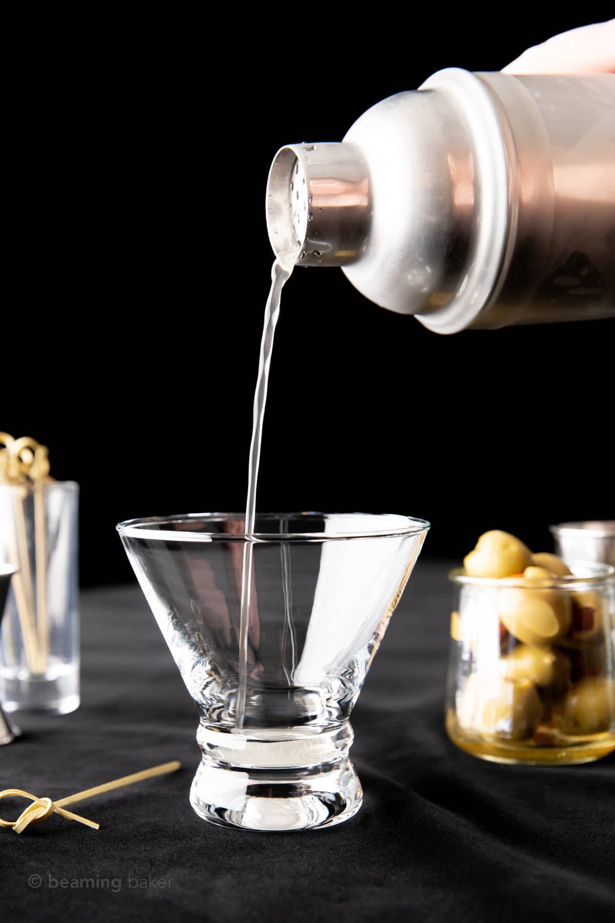 A hand pouring liquid from a stainless steel cocktail shaker into a martini glass